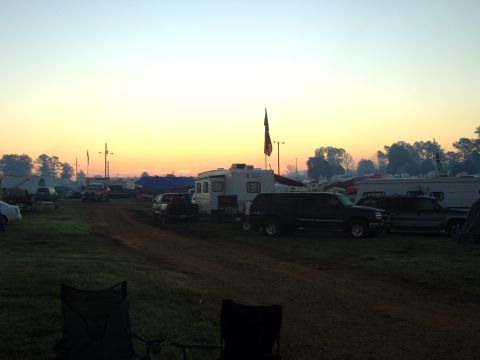 2007 Ford Mustang - A campsite scene unfolds at sunrise, with RVs and various vehicles parked amidst a grassy and dirt terrain, while two chairs sit in the for…