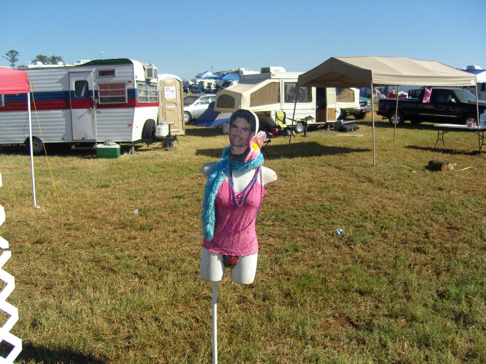 Mannequin dressed in pink top and blue scarf stands in a grassy field with campers and tents in background