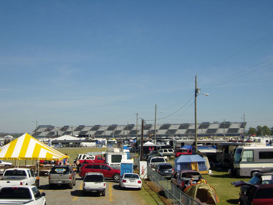Parking lot with many cars and RVs, yellow and white tent in foreground, racetrack grandstands in background under a clear blue sky
