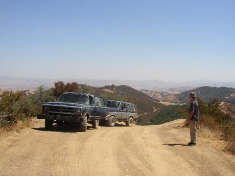 1989 Ford Bronco - Two SUVs are parked on a dirt road with a man standing to the side, overlooking a scenic valley.