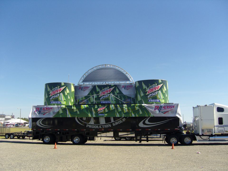 Stage and advertising setup on a trailer with a semi-truck attached, under a clear blue sky