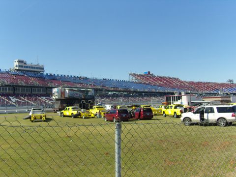 2007 Ford Mustang - Numerous yellow trucks and red vans are parked on the infield grass at a racetrack, with spectators in the stands.