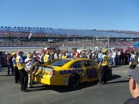 2007 Ford Mustang - A yellow race car with "KOMATSU" and "09" logos is on the track with crew members and spectators around.