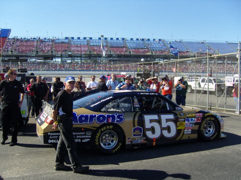 2007 Ford Mustang - A race car with "Aaron's" livery is parked near a grandstand with spectators.