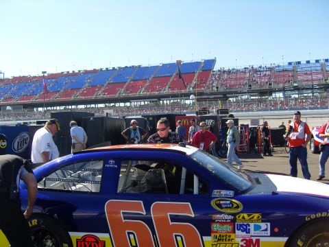 2007 Ford Mustang - Race car #66 is parked in the pit lane with crew members around it and grandstands in the background.