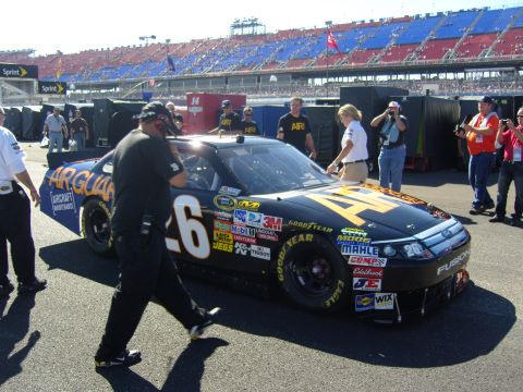 2007 Ford Mustang - A race car with numerous sponsor decals is parked on the track, with people and grandstands visible.