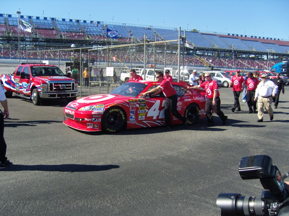 Red NASCAR race car with number 42 and Target logos, crew members pushing it, red Ford truck nearby, grandstands in background