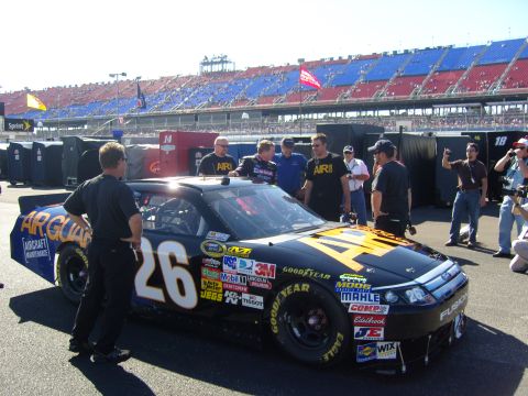 2007 Ford Mustang - A black race car with "AIRGUARD" livery and the number 26 is parked at a racetrack, with people gathered around.