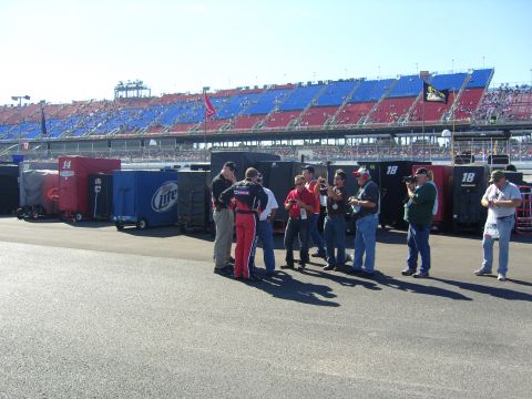 2007 Ford Mustang - A group of people, some in racing attire, stand in a pit area with a large grandstand filled with spectators behind them.