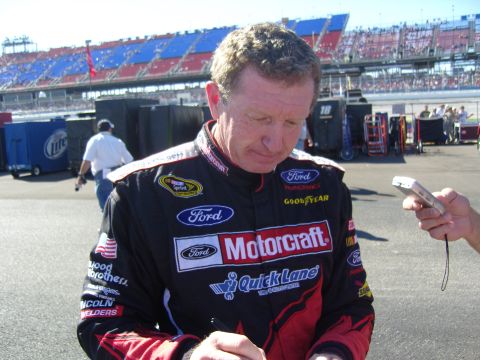2007 Ford Mustang - A NASCAR driver in a racing suit with sponsor logos is signing an autograph at a race track.