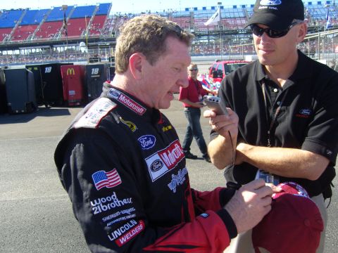 2007 Ford Mustang - A race car driver is interviewed at a track, with grandstands and pit areas visible in the background.
