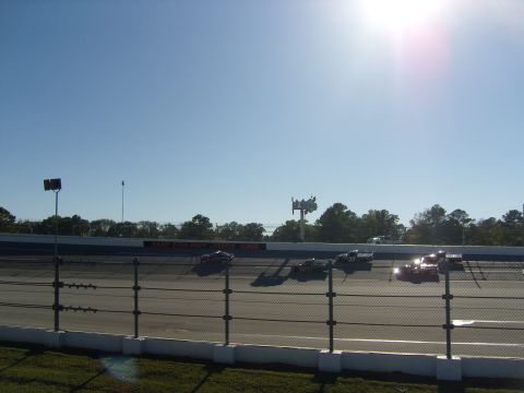 2007 Ford Mustang - Multiple race cars are seen on a track during a sunny day, with trees and a light tower in the background.