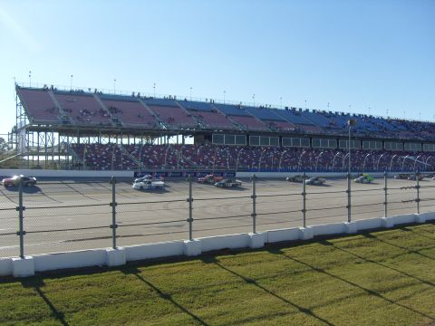 2007 Ford Mustang - Multiple stock cars compete on a race track with a full grandstand under a clear blue sky.