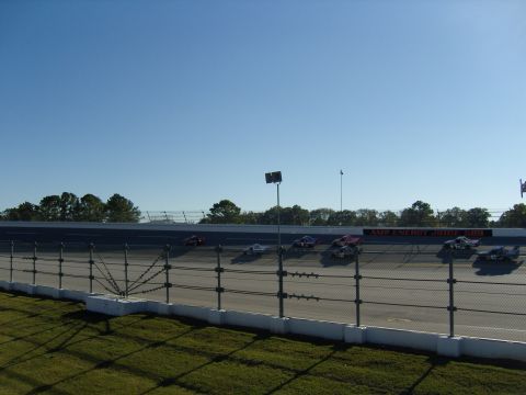 2007 Ford Mustang - Several stock cars are racing on a track under a clear blue sky, viewed from behind a fence.
