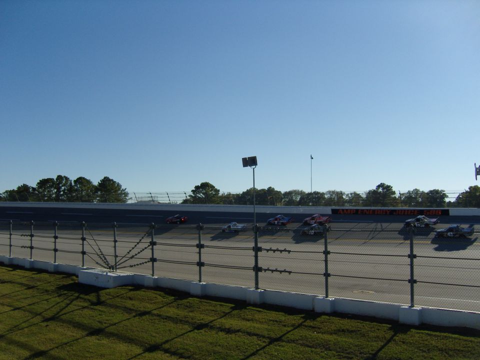stock cars racing on a track with a fence in the foreground and trees in the background