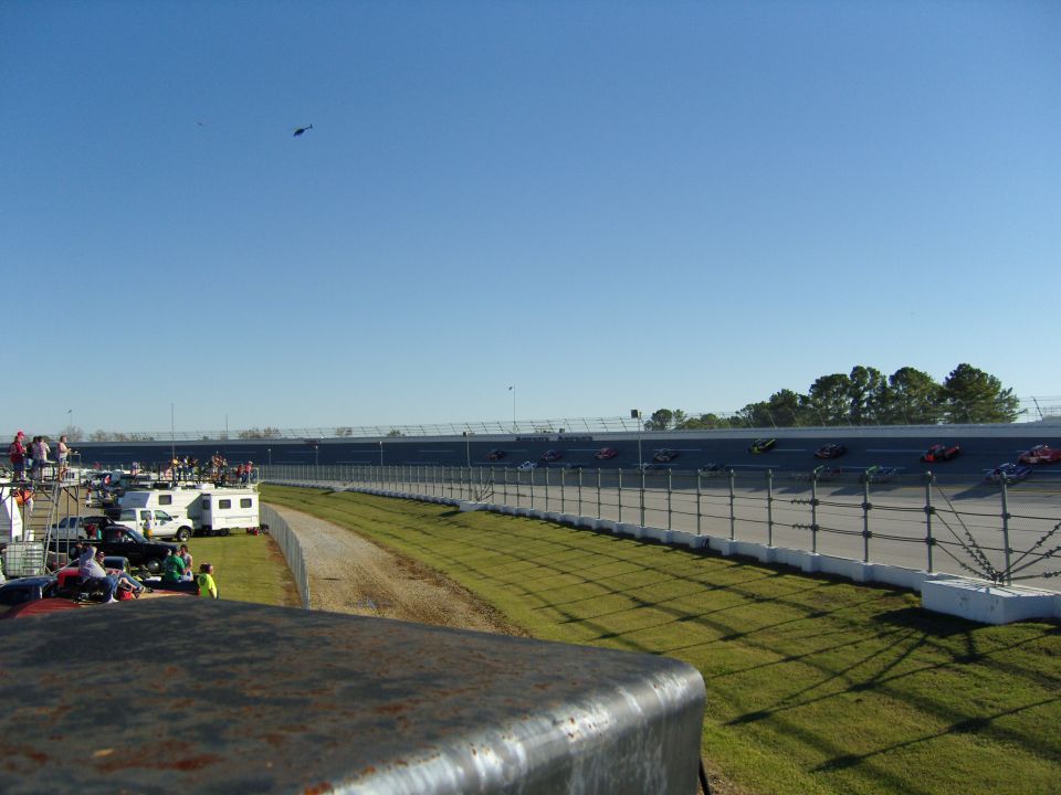 Race cars on a track with spectators and RVs in the background under a clear blue sky