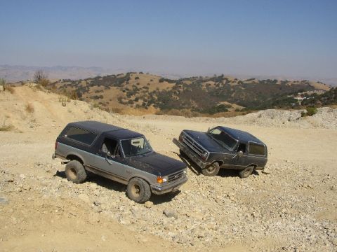1989 Ford Bronco - Two 1989 Ford Broncos are parked on a dirt hill under a clear blue sky.