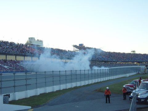 2007 Ford Mustang - Smoke billows across a race track in front of a large crowd in the stands, with several vehicles visible in the foreground.