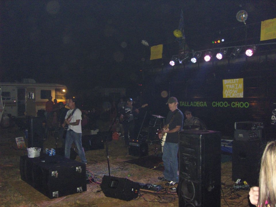 Band playing on an outdoor stage at night with RVs in the background. Text on the stage reads 'TALLADEGA CHOO-CHOO'