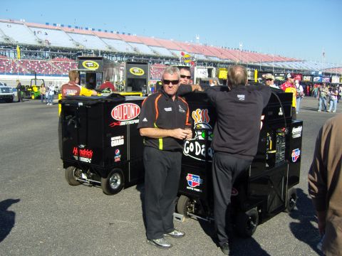 2007 Ford Mustang - Race personnel stand by equipment at a speedway, with grandstands visible in the background.