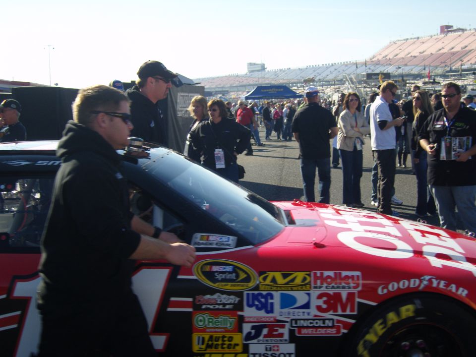 Red NASCAR race car with numerous sponsor decals, including Goodyear and 3M. People are visible in the background near a race track
