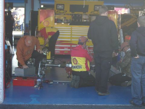 2007 Ford Mustang - Mechanics are gathered around a vehicle chassis in a garage, with a large yellow toolbox in the background.