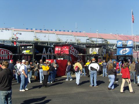 2007 Ford Mustang - A crowd of people stand in front of various sponsor trailers at a race track event.