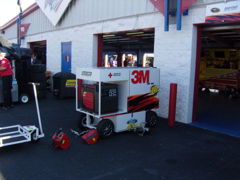 2007 Ford Mustang - Generator and equipment cart with 3M and American Red Cross logos is parked outside a garage at a race track.