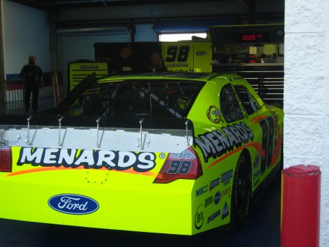 2007 Ford Mustang - A bright yellow race car with "MENARDS" branding is parked inside a garage bay.