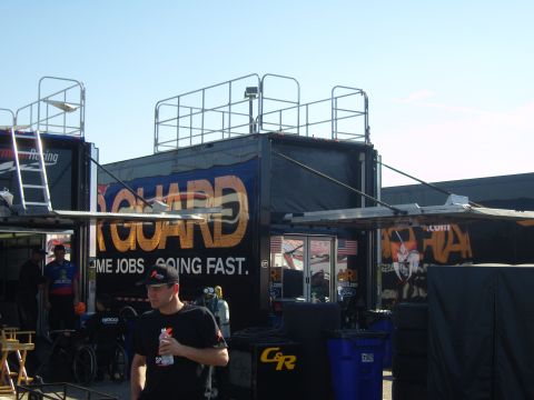 2007 Ford Mustang - Pit area scene with large support trucks and crew members under a clear sky.