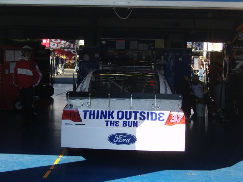 2007 Ford Mustang - A race car is parked in a garage, showing its rear with sponsor decals and the Ford logo.