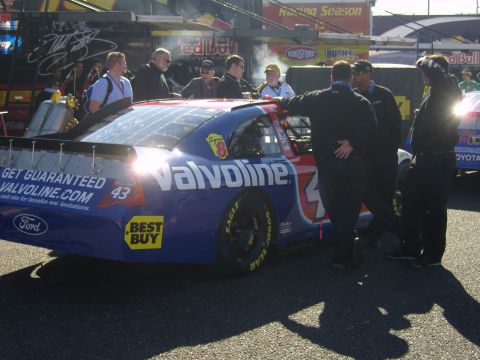 2007 Ford Mustang - A race car with sponsor logos is parked in a pit area with crew members around it.