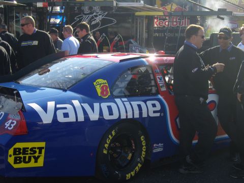 2007 Ford Mustang - A race car with sponsor logos is parked in a pit area with crew members around it.