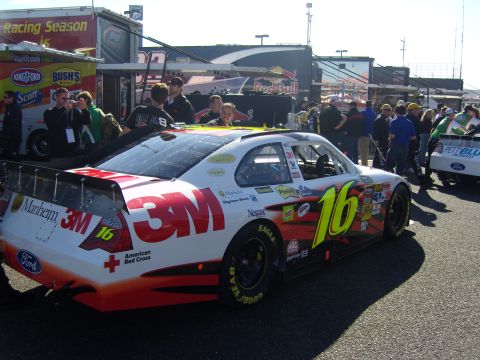 2007 Ford Mustang - A NASCAR race car with sponsor decals is parked at the track, with other vehicles and people in the background.