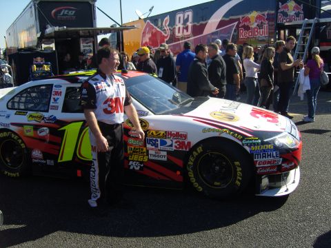 2007 Ford Mustang - A race car with a man in a racing suit stands next to it, with a large Red Bull branded trailer in the background.