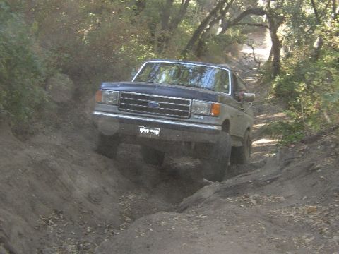 1989 Ford Bronco - This Ford Bronco navigates a challenging off-road trail surrounded by trees.