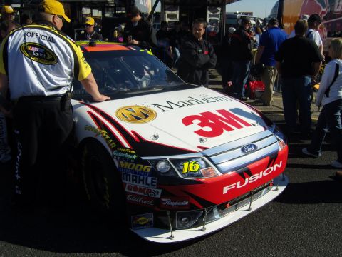 2007 Ford Mustang - A NASCAR race car with various sponsor decals is parked on asphalt with people around.