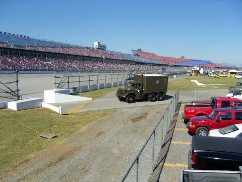 2007 Ford Mustang - Military truck on track infield with grandstands and parked vehicles visible.