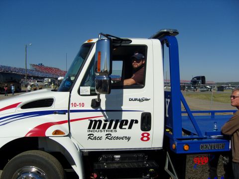 2007 Ford Mustang - A tow truck with "Miller Industries Race Recovery" branding is parked at a venue with grandstands in the background.
