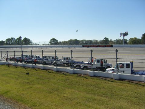 2007 Ford Mustang - Multiple tow trucks are parked in a line along the side of a race track under a clear blue sky.