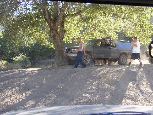Muddy dark two-door SUV parked on dirt with men standing nearby under a tree