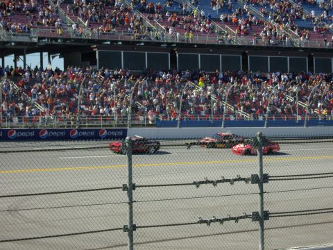 2007 Ford Mustang - Race cars speed around the track with spectators filling the stands above.