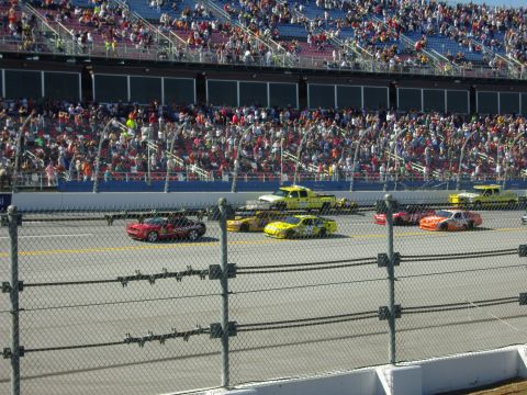 2007 Ford Mustang - Multiple race cars compete on a track in front of a large crowd in the grandstands.