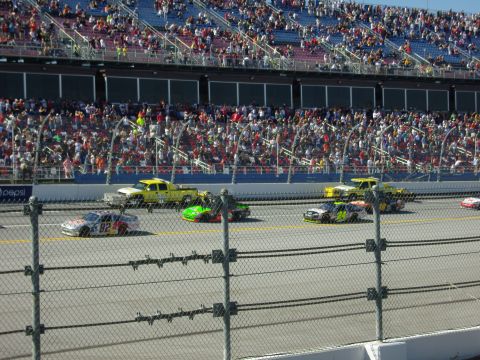 2007 Ford Mustang - Multiple race cars compete on a track in front of a large crowd in the grandstands.
