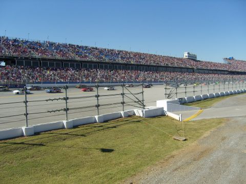 2007 Ford Mustang - Multiple race cars compete on a track in front of a packed grandstand under a clear blue sky.