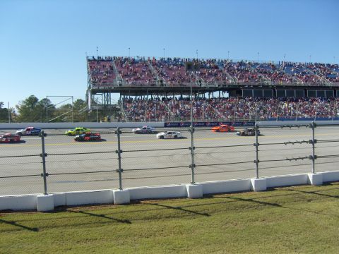 2007 Ford Mustang - Multiple stock cars are racing on a track in front of a large grandstand filled with people.