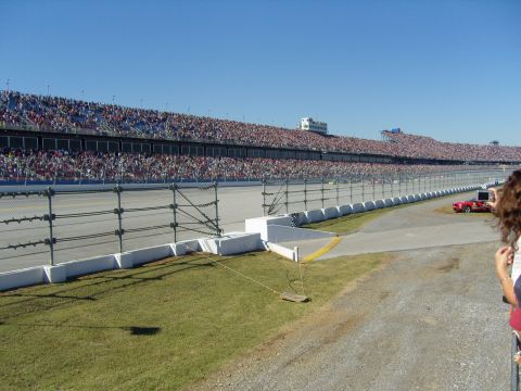 2007 Ford Mustang - A red car is visible on the racetrack, with a large crowd in the grandstands under a clear blue sky.