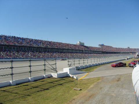 2007 Ford Mustang - A red 2007 Ford Mustang is visible on the side of a racetrack, with a large crowd in the stands and a helicopter in the sky.