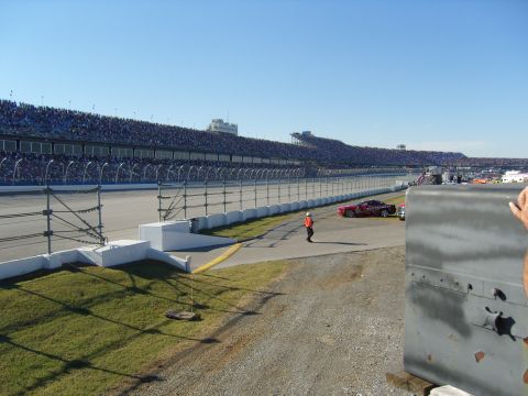 2007 Ford Mustang - A red race car is parked on the track with a person in an orange vest nearby, under a clear blue sky.
