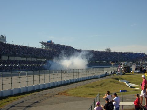 2007 Ford Mustang - A race car creates a large smoke cloud on the track as spectators fill the grandstands under a clear blue sky.